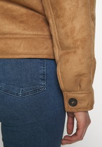 Tan faux suedejacka med en smidig textur, med knappstängning vid ärmen. Matchad med mörkblå denimjeans.