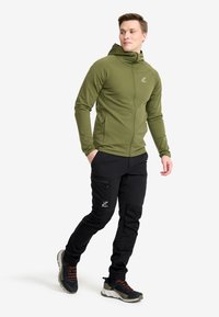 Olive green zip-up hoodie with a fitted design and hood. Paired with black cargo pants featuring zip pockets and rugged black shoes.
