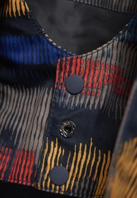 Tessuto a fantasia con strisce blu, rosse e gialle, caratterizzato da un design a spina di pesce testurizzato. Bottoni neri e un evidente dettaglio di chiusura a scatto.