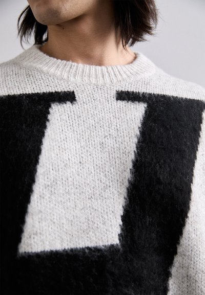 Kötött pulóver krém és fekete textúrával, kiemelkedő fekete "L" mintával. A nyakkivágása bordás és kerek fazonú.