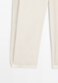 Nedre delen av beige byxor med rakt bensnitt med synliga sidsömmar och sydda fållar på vit bakgrund.