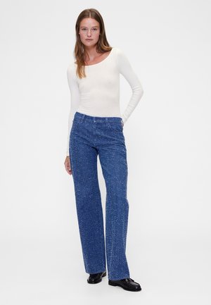 Dugotrajna kremasta majica rebrastog uzorka i plave hrapave široke traperice; majica ima okrugli izrez. Tamne cipele upotpunjuju odjeću.