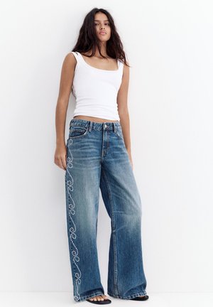 Ung kvinde iført hvid ærmeløs top og vidde bukser med dekorative hvide nitter langs sidesømmen, stående op ad hvid væg.