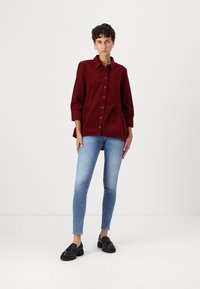 Burgunderfarget skjorte med strukturert silhuett, parret med tettsittende lyseblå jeans og svarte loafers. Glatt stofftekstur.