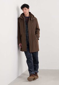 Bruine waterdichte jas met capuchon, rits aan de voorkant en trekkoorden. Draag deze over een gestreept shirt, gecombineerd met corduroy broeken en bruine schoenen.