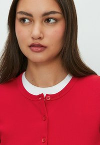 Jonge vrouw met lang bruin haar, gekleed in een rode blouse over een wit shirt, kijkt naar links tegen een effen achtergrond.