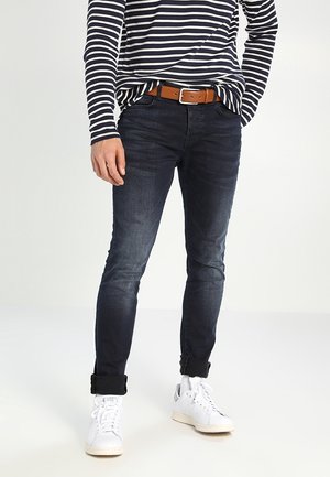 Donkerblauwe jeans met een slim fit, vervaagde details en opgerolde mouwen. Gecombineerd met een gestreept shirt met lange mouwen en witte sneakers. Bruine riem.
