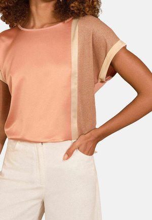 Femme portant un top pêche à manches courtes avec une bande verticale beige brillante, associé à un pantalon taille haute écru, main dans la poche.
