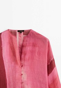 Chemise en lin rose avec un col en V, dotée d'une fermeture à boutons sur le devant et de variations de couleur subtiles pour la texture. Conception fluide avec des manches longues.