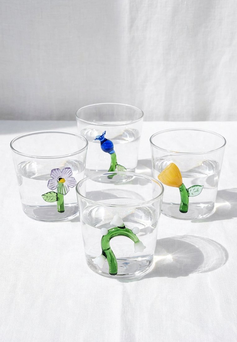 Cztery przezroczyste szklanki wypełnione wodą, każda zawierająca mały zielony szklany łodyg z kolorowymi kwiatowymi lub roślinnymi kształtami w środku.