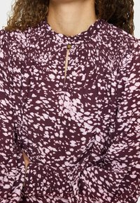 Donna che indossa una blusa a maniche lunghe di colore viola con macchie astratte rosa e una piccola chiusura a bottone vicino al colletto.