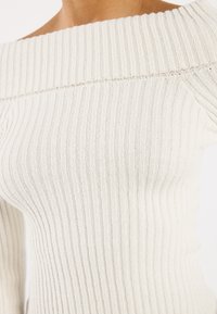 Rebrast off-shoulder top v svetlo bežovi, ki se ponaša s strukturiranim pletenim vzorcem in oprijetim silhueto, ki izpostavlja ovratnik in robove rokavov.
