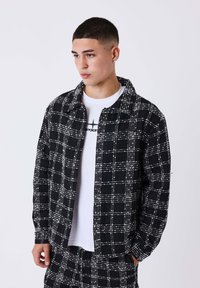 Jonge man met kort haar, gekleed in een zwart-wit geruit jasje en bijpassende broek boven een witte graphic t-shirt, kijkend opzij.