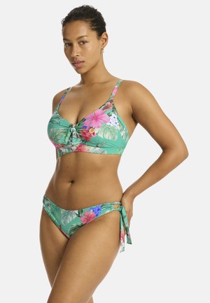 Kobieta ubrana w zielony kwiatowy bikini z różowymi, czerwonymi i niebieskimi kwiatami, pozująca z jedną ręką na biodrze, neutralne tło.