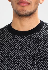 Czarny sweter z wzorem w zygzak w odcieniach szarości, z żeberkowym dekoltem i miękką teksturą. Zbliżenie na górny obszar klatki piersiowej.