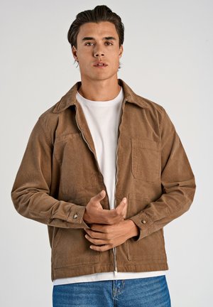 Chaqueta de pana marrón con cuello de camisa, dos bolsillos delanteros y cremallera. Llevada sobre una camisa blanca de textura suave.
