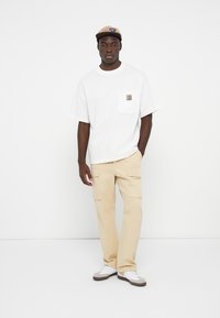 White oversized t-shirt with a chest pocket logo, paired with beige cargo pants featuring side pockets, and white sneakers with gray accents.