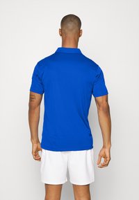 Polo a maniche corte da uomo in tessuto blu brillante. Presenta un colletto classico ed è abbinato a pantaloni corti bianchi, mostrando una trama liscia.