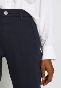 Tummansiniset housut, joissa on suora leikkaus, etutaskut ja hopeinen nappikiinnitys, yhdistettynä valkoiseen kauluspaitaan.