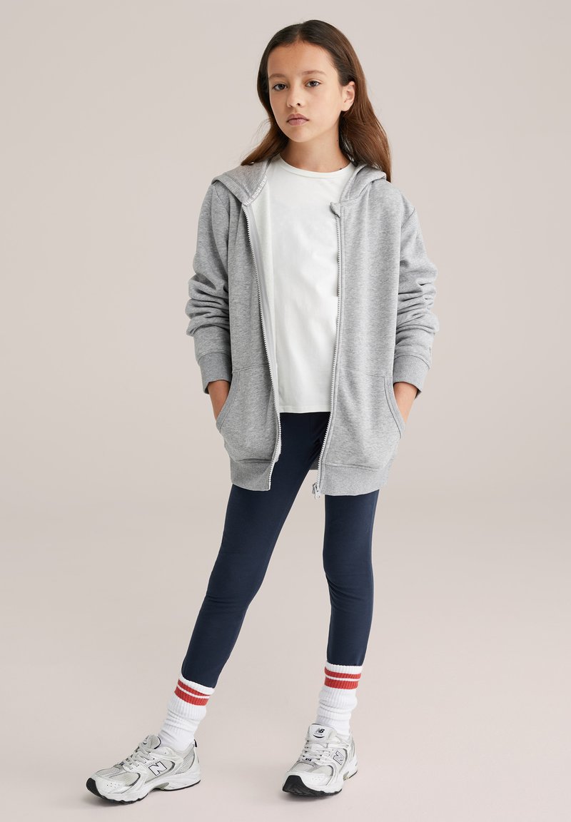 Szürke cipzáras pulóver nagy kapucnival, fehér pólóval és sötétkék leggingssel párosítva. Az öltözéket piros csíkos fehér zokni és szürke sportcipő teszi teljessé.