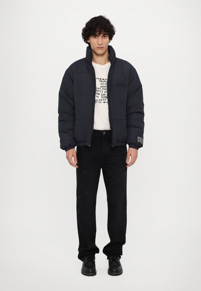 Doudoune marine avec un col montant et des coutures visibles, associée à un pantalon en denim noir avec des motifs de croix brodés. Chaussures noires.