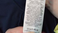 Textilápolási címke egy szöveten. Többnyelvű utasításokat tartalmaz, fehér szöveg világosszürke háttéren, az alján ápolási szimbólumokkal.