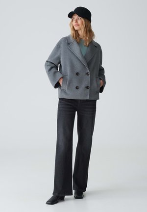 Gray, double-breasted coat with wide sleeves, black buttons, and two pockets, worn over a teal top, paired with dark jeans and black boots.