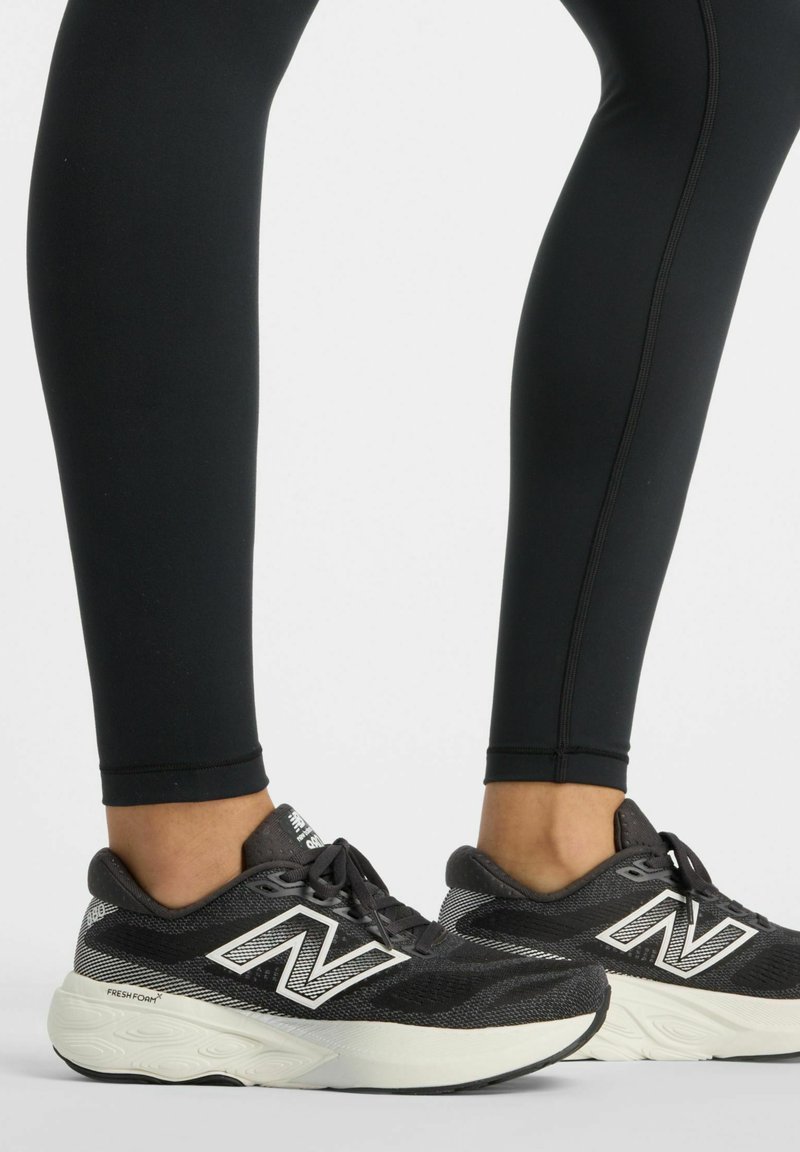 Leggings noirs associés à des baskets New Balance noires et blanches, dotées d'une tige en mesh texturé, d'une semelle rembourrée et d'un logo "N" bien visible.