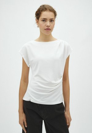 Jeune femme aux cheveux bouclés portant un haut blanc à manches courtes et un pantalon noir, debout devant un fond clair uni.