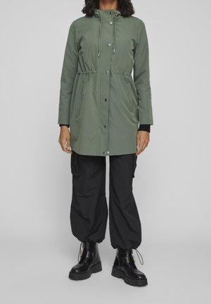 Mujer con impermeable verde con capucha, pantalones cargo negros con puños elásticos y botas negras gruesas de tobillo, de pie sobre un fondo gris.