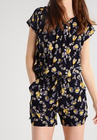 Combinaison florale noire avec des fleurs jaunes et grises. Manches courtes, taille cintrée et poches. Fabriquée en tissu léger avec une texture douce.