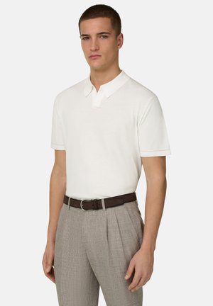 Mężczyzna w białej koszulce polo z krótkim rękawem wpuszczonej w beżowe spodnie z fałdami, z ciemnobrązowym skórzanym paskiem, stojący na jednolitym tle.