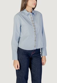 Camicia azzurro chiaro con colletto abbottonato, caratterizzata da una linea decorativa di accenti floreali argentati lungo la parte anteriore, abbinata a pantaloni in denim scuro.