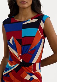 Mujer que lleva un vestido sin mangas con estampado geométrico en rojo, azul, negro, naranja y beige, que presenta un detalle de nudo en la parte delantera.