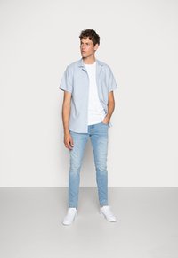 Helles blaues kurzärmliges Hemd, darunter ein weißes T-Shirt mit Rundhalsausschnitt, kombiniert mit hellblauen Jeans und weißen Sneakers, stehend vor einem neutralen Hintergrund.