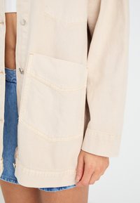 Beige denimjakke med en frontlomme, knaplukning og synlige stiksømme. Stoffet virker blødt med en let tekstureret overflade.
