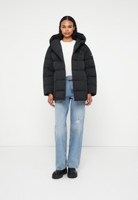 Jachetă puffer neagră cu glugă, având un design pătrățos, textură matlasată și o buzunar cu fermoar pe brațul stâng. Purtați peste o cămașă albă și blugi blue ripped.