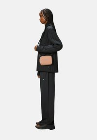 Giacca nera trapuntata, pantaloni neri larghi e borsa a tracolla marrone. Il modello indossa sandali neri. Design minimalista, look monocromatico.