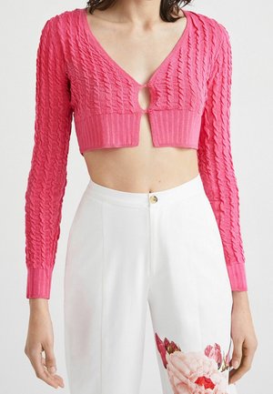 Femme portant un cardigan court en maille rose vif avec un détail bouton en forme d'œillet, associé à un pantalon blanc taille haute avec imprimé floral.