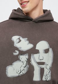 Felpa con cappuccio marrone con stampe grafiche di due volti, uno con i capelli e l'altro con le mani sulle guance, in tonalità bianche e grigie. Tessuto morbido e strutturato.