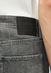 Κοντινό πλάνο της τσέπης από γκρι τζιν με την μαύρη ετικέτα Jack & Jones και μέρος ενός λευκού πουκάμισου και χεριού ορατά.