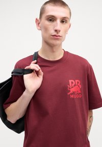 Maroon cotton t-shirt with a crew neck, featuring a red graphic of a bull and text on the chest. Model holds a black bag strap.