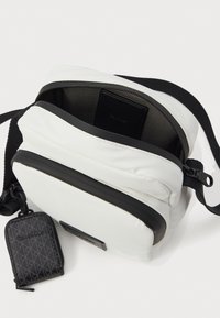 Λευκή τσάντα crossbody με μαύρο φερμουάρ και λουρί, ανοιγμένη εν μέρει που δείχνει μια μαύρη θήκη μέσα, καθώς και ένα μικρό μαύρο pouch της Calvin Klein συνδεδεμένο.