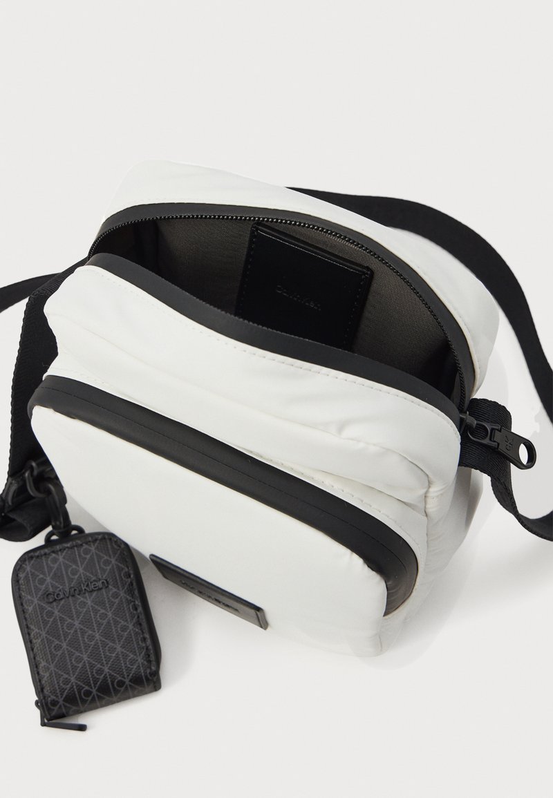 Bolso cruzado blanco con cremallera y correa negra, parcialmente abierto mostrando una cartera negra en el interior y un pequeño estuche negro de Calvin Klein adjunto.
