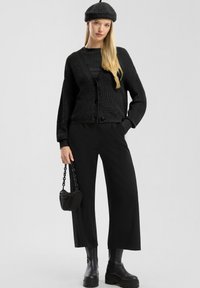 Cardigan en tricot noir avec un col en V, fermeture à boutons, associé à un pantalon noir ample et des bottes noires imposantes. Les accessoires comprennent un petit sac et un béret.