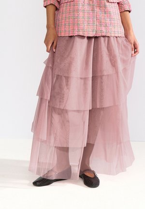 Personne portant une jupe rose transparente à plusieurs couches, une veste rose et blanche texturée, et des ballerines tissées noires debout sur un sol blanc.