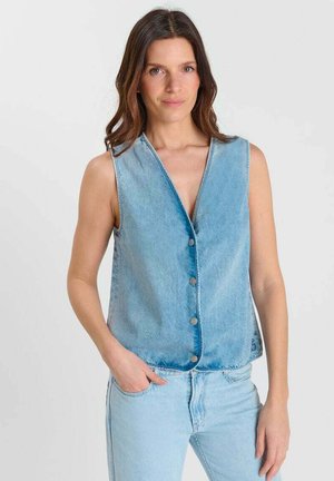 Vrouw met lang donker haar, gekleed in een lichtblauwe denim spijkervest zonder mouwen met knopen en bijpassende denim spijkerbroek, staand met één hand in haar zak.