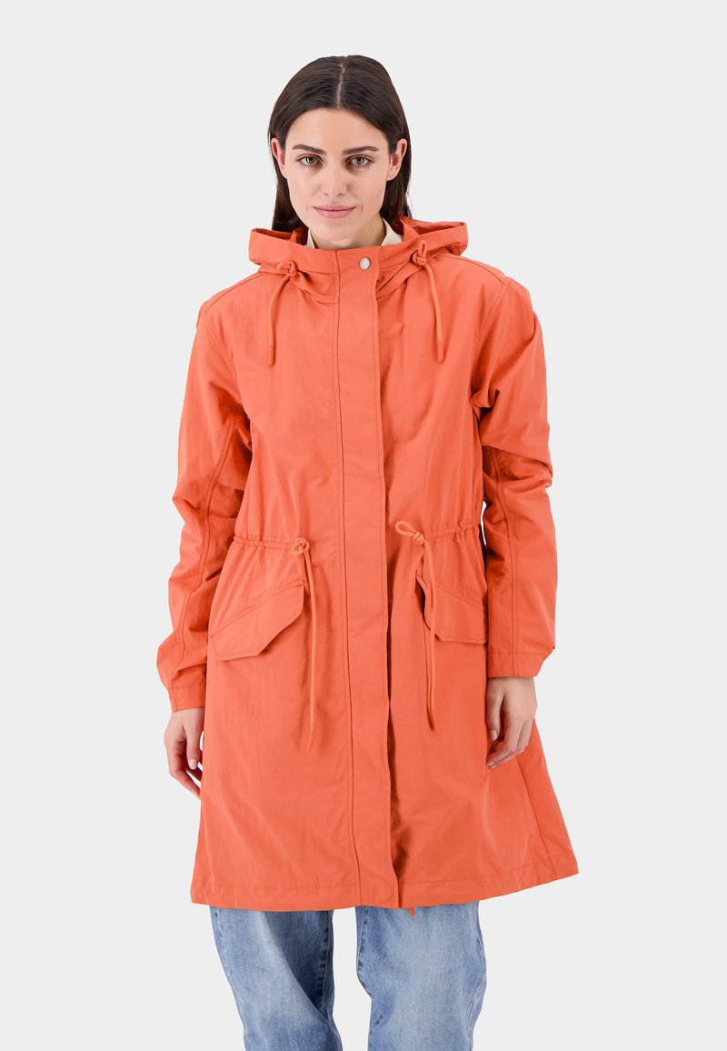 Vrouw met steil, donker haar die een oranje hoodiejas met trekkoorden en grote voorzakken draagt over lichtblauwe jeans.