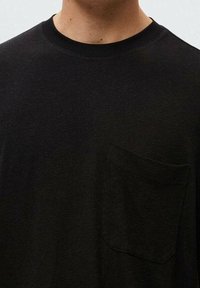 Person iført en ensfarvet sort T-shirt med rund hals og en lille brystlomme på venstre side, mod en lys baggrund.