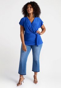 Top avvolgente blu in tessuto morbido con maniche corte, legato in vita. Abbinato a jeans croppati blu chiaro con orli sfrangiati. Tacchi a spillo argentati.